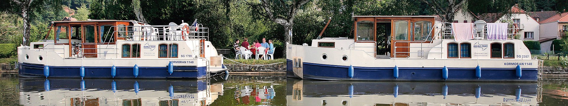 Zwei Kormoran-Hausboote haben am Ufer angelegt. Die Passagiere habe es sich an Land gemütlich gemacht und sitzen an Tischen und schwatzen.